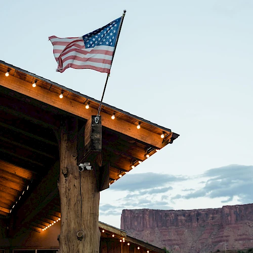 An American flag on a pole above a wooden porch with string lights, set against a canyon backdrop.