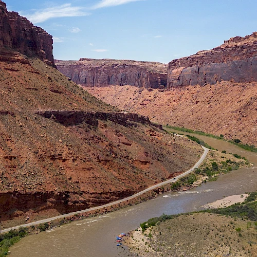 A muddy river winds through a canyon with reddish cliffs and green shrubs, a dirt road tracing along the riverbank in a vast valley.