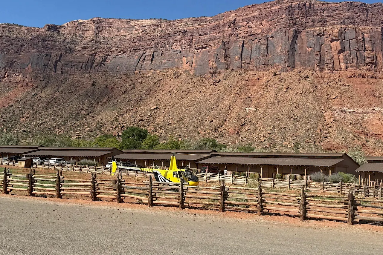 A yellow rescue helicopter sits near a desert lodge, backed by red canyon cliffs and a fenced road.