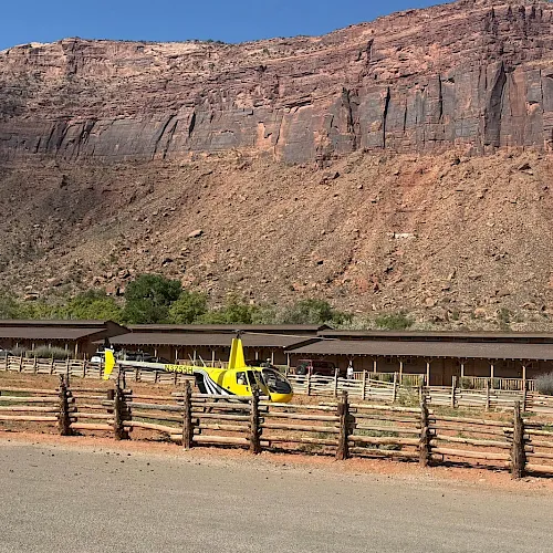 A yellow rescue helicopter sits near a desert lodge, backed by red canyon cliffs and a fenced road.
