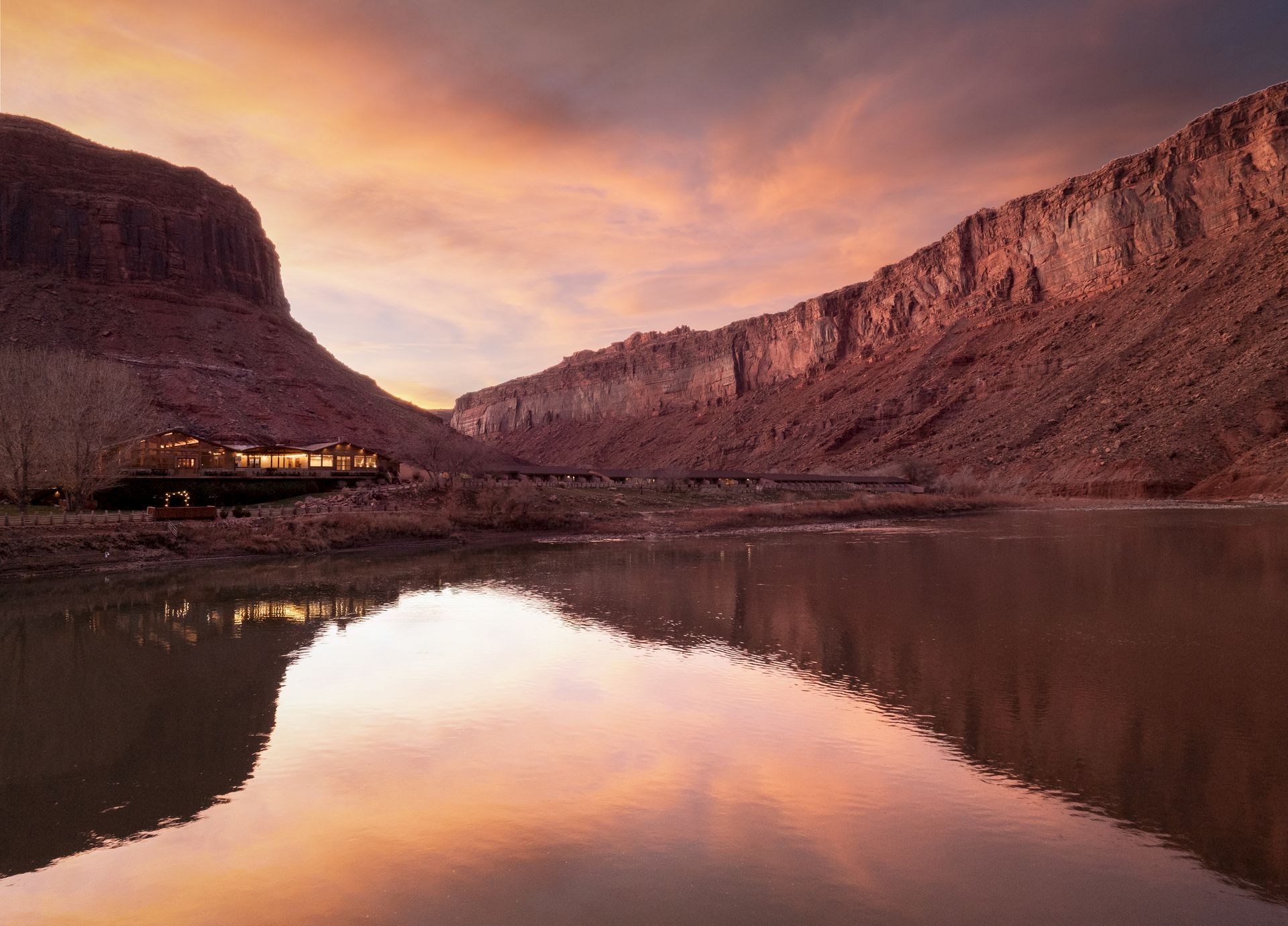 A tranquil river mirrors a dramatic canyon at sunset, with warm clouds painting the sky and a small settlement glowing on the left shore.