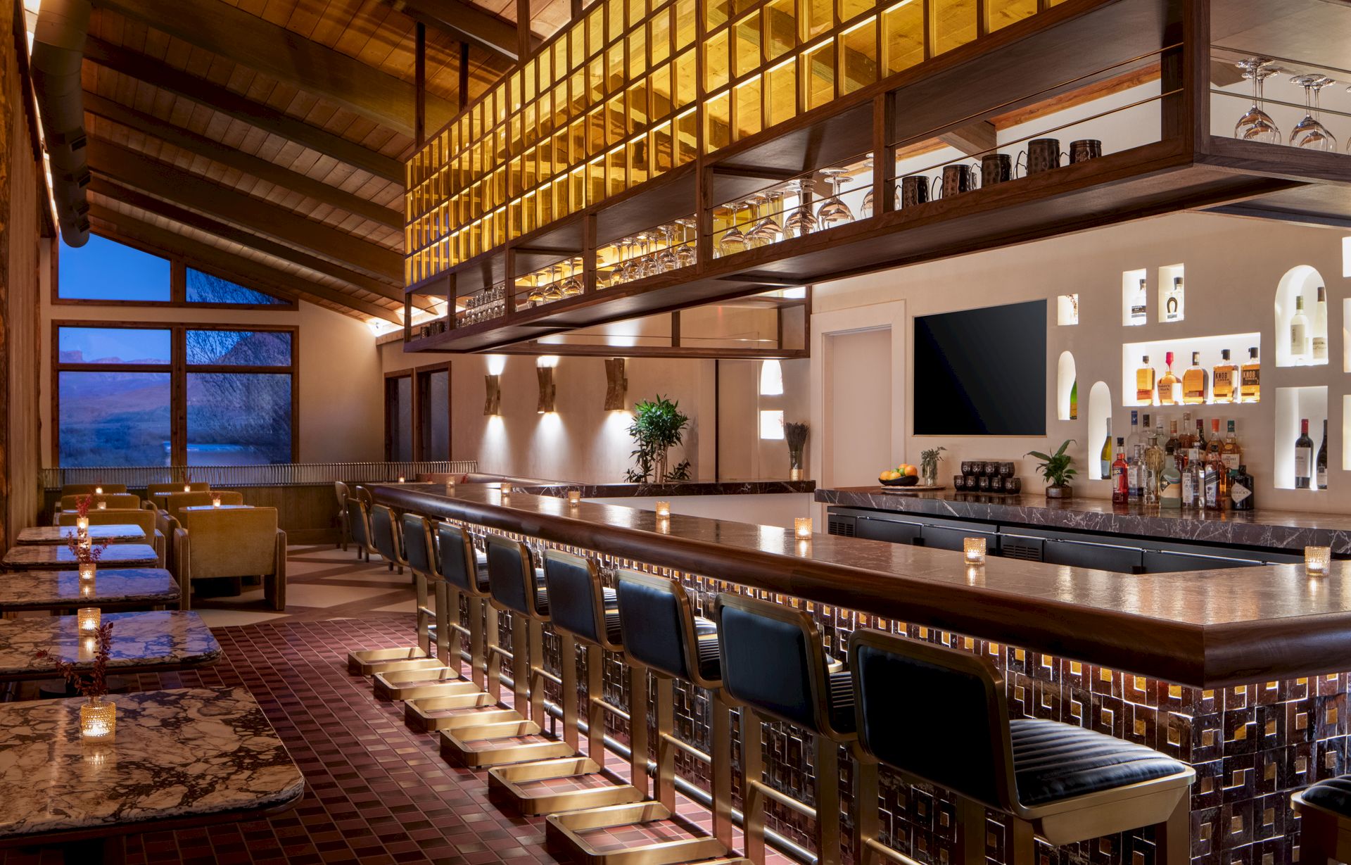 A stylish, warmly lit bar with a long wooden counter, cushioned stools, wine shelves overhead, and a cozy lounge area by the windows, inviting and chic.