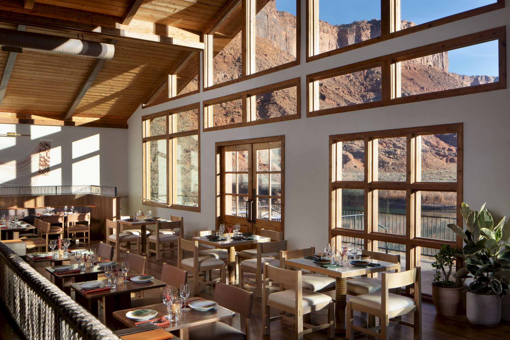 A bright restaurant with wooden tables and chairs, large windows showing a canyon view, warm wood beams, and plant decor, cozy dining space.