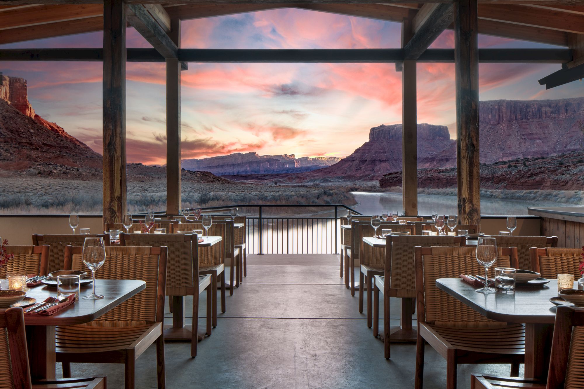 A cozy outdoor dining area with wicker chairs and tables set for dinner, facing a dramatic desert canyon sunset over a tranquil lake.