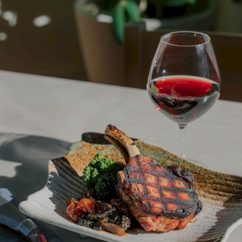 A plated steak with grilled marks, greens, bread, a wine glass of red wine, and a bottle opener on a napkin nearby, set on a table.