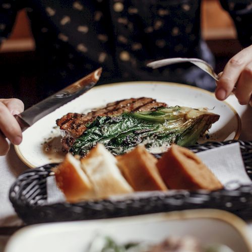 Two people share a plated asparagus-tish steak meal, cutting with knives as bread sits in a basket nearby, dinner setting looks cozy.