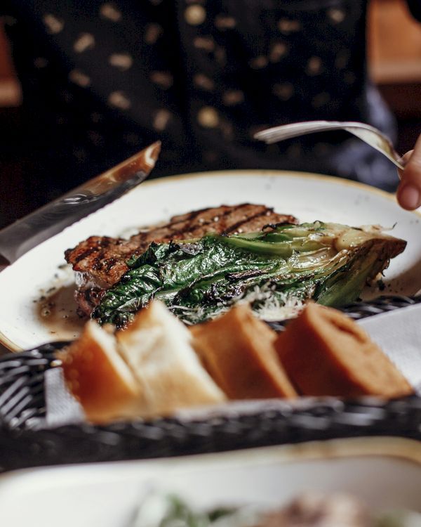 Two people share a plated asparagus-tish steak meal, cutting with knives as bread sits in a basket nearby, dinner setting looks cozy.