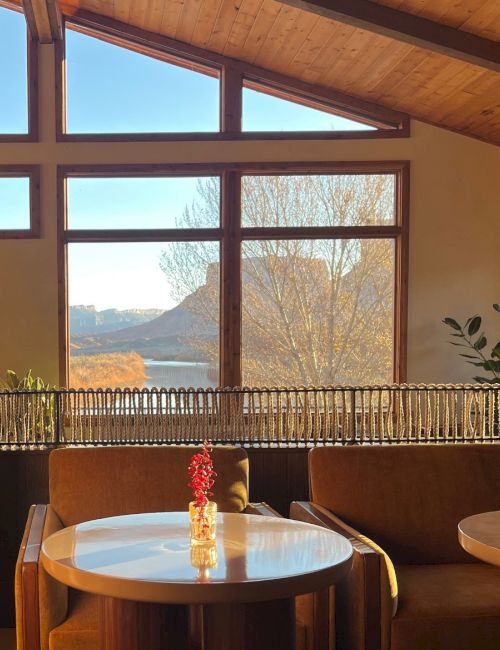 Cozy indoor seating with a round table, brown chairs, a vase of flowers, and a large window framing a scenic lake and bare trees outside, under a wooden ceiling.