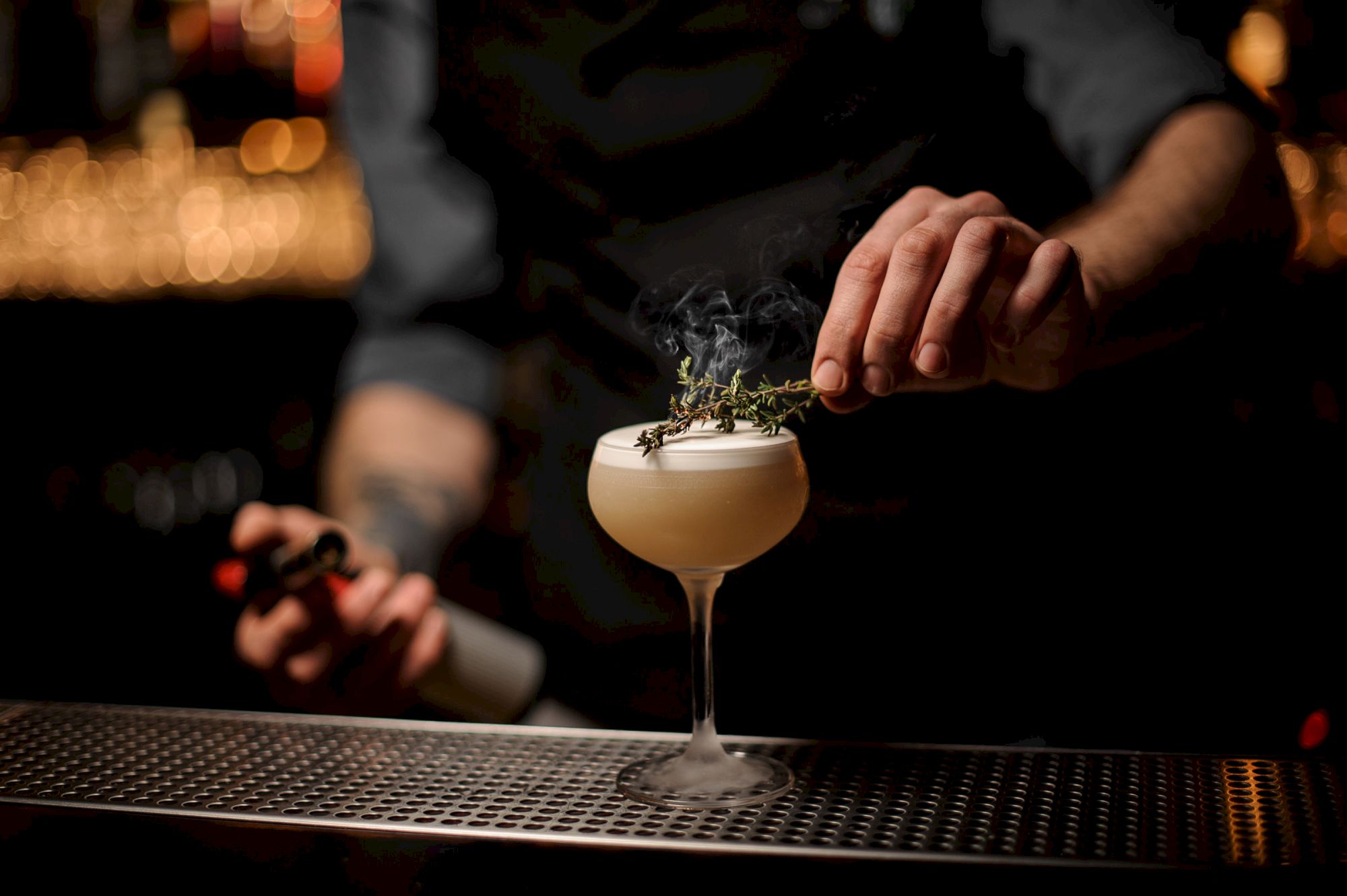 A bartender tops a fancy cocktail with herbs over a frothy, pale drink on a bar counter, smoke or garnish drifting above the glass.