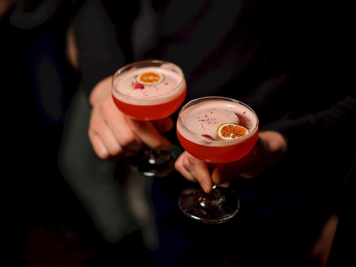 Two people clink champagne coupe glasses with pink cocktails, garnished, in a dark setting. end the sentence.