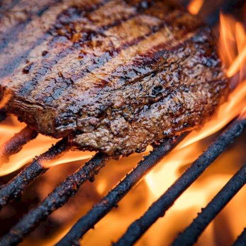 A close-up of glowing charcoal embers and sizzling grill grate over a fire, with orange flames licking the metal.
