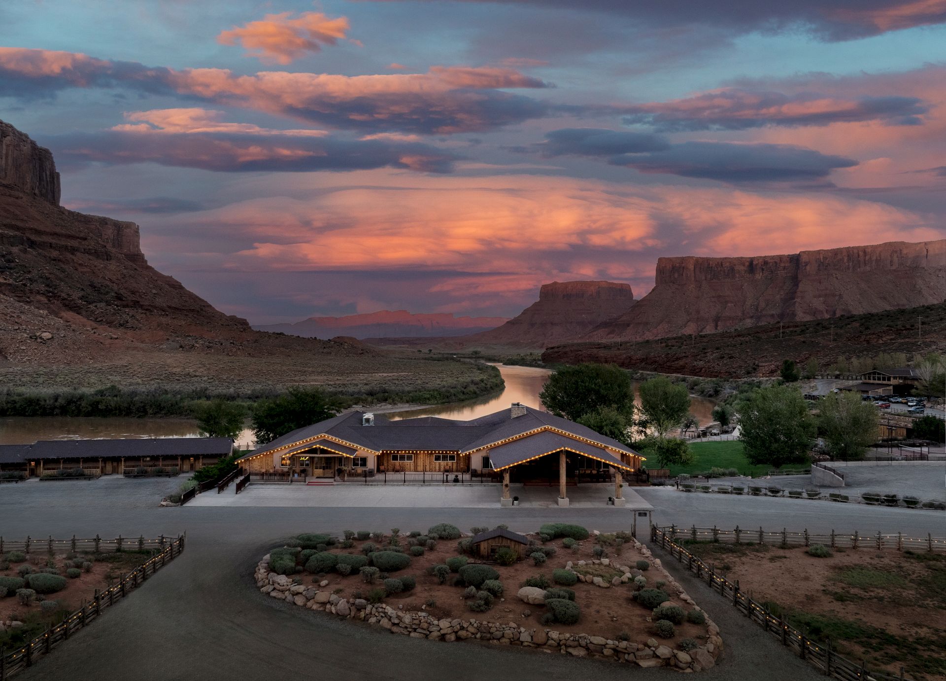 Sunset over a canyon lodge with a winding road, rugged cliffs, and a circular landscaped front yard in a desert valley.