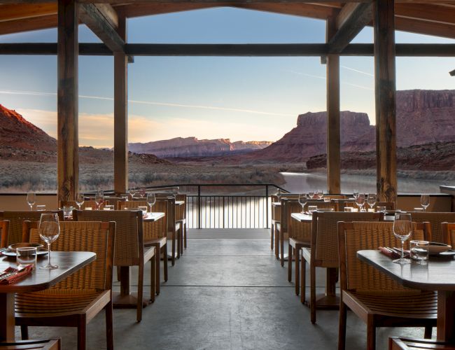 A stylish outdoor restaurant with wicker chairs, tables set for dinner, overlooking a dramatic desert canyon at sunset, under a wooden pergola.