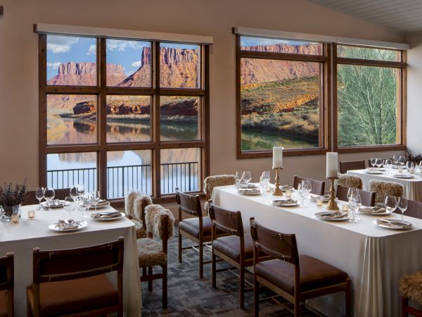 A dining room with white-clothed tables, elegant place settings, and large windows overlooking a desert canyon landscape, warm lighting, and wooden chairs.
