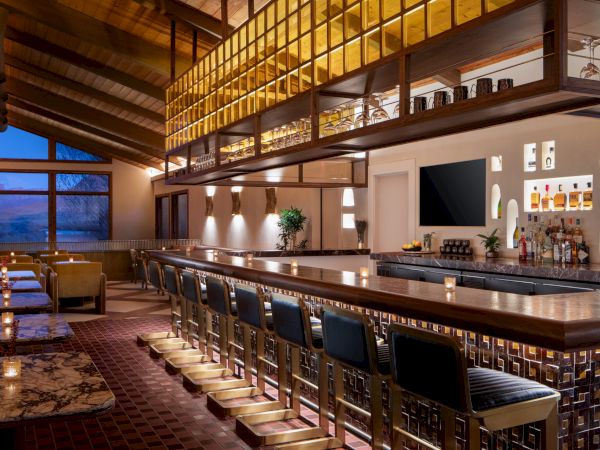 A stylish bar scene with a long illuminated counter, high stools, hanging glass rack, warm wood tones, and coastal sunset view through the windows.