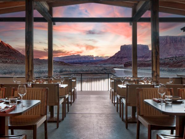 An open-air restaurant with wicker chairs and tables set for dining, facing a sunset over a canyon landscape with pillars framing the view.
