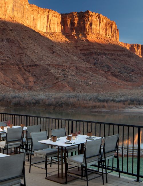 Outdoor dining terrace overlooking a calm river and towering red rock mesas at sunset, with tables set and a railing along the edge.
