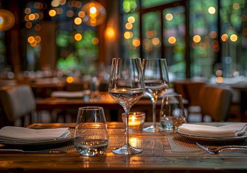 A dimly lit restaurant table set with wine glasses, candles, plates, and cutlery; warm bokeh lights glow in the background.