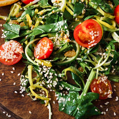 Fresh greens, cherry tomatoes, and spiralized zucchini noodles tossed with sesame seeds and herbs on a wooden plate. End.