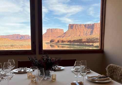 A dining table set for two with white plates, silverware, glassware, and a centerpiece, overlooking a desert canyon through large windows, at sunset.