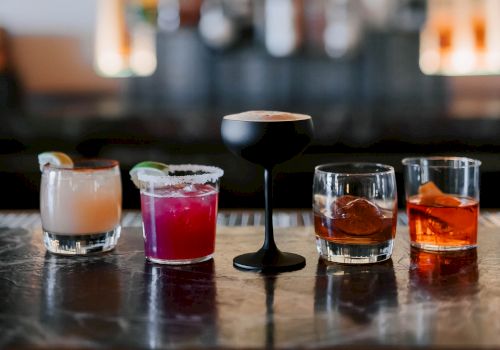 A row of colorful cocktails on a bar counter, from left to right: creamy pink, purple, a tall black-stemmed glass, and amber-toned whiskey drinks, all ready to toast.