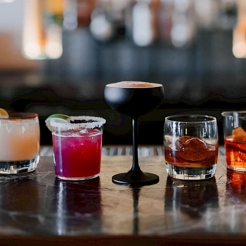 A row of colorful cocktails on a bar counter, from left to right: creamy pink, purple, a tall black-stemmed glass, and amber-toned whiskey drinks, all ready to toast.