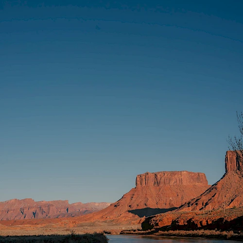 A tranquil desert river winds through red rock formations under a clear blue sky, reflecting the landscape in its calm waters.