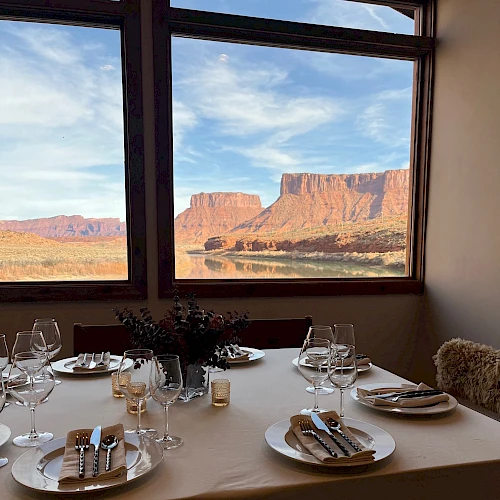 A dining table set for two with white plates, silverware, glassware, and a centerpiece, overlooking a desert canyon through large windows, at sunset.