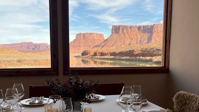 A dining table set for two with white plates, silverware, glassware, and a centerpiece, overlooking a desert canyon through large windows, at sunset.