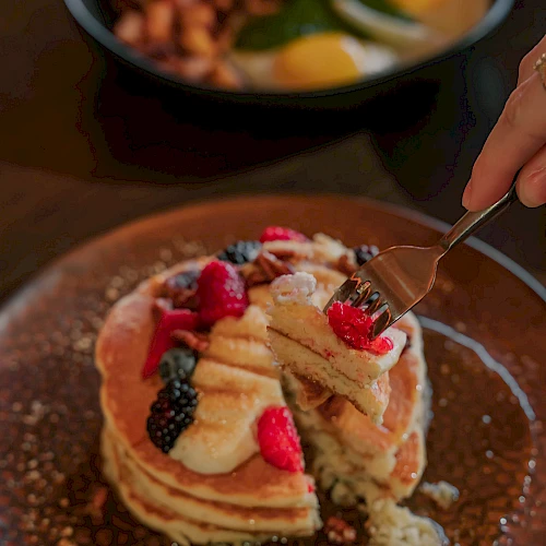 A stack of fluffy pancakes topped with sliced bananas, berries, and dollops of whipped cream, with syrup pooling around the plate.