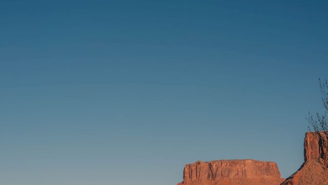 A calm river wends through a canyon with red rock formations and a clear blue sky above, their reflections shimmering on the water&rsquo;s surface.
