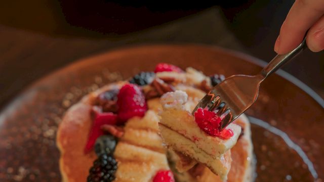 A stacked pancake dessert topped with banana slices, raspberries, blackberries, and other berries, with a spoon about to take a bite.