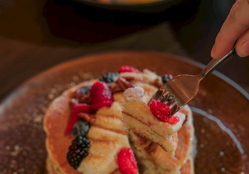 A stack of fluffy pancakes topped with sliced bananas, raspberries, blackberries, and a drizzle of syrup on a brown plate, with a fork about to cut in.