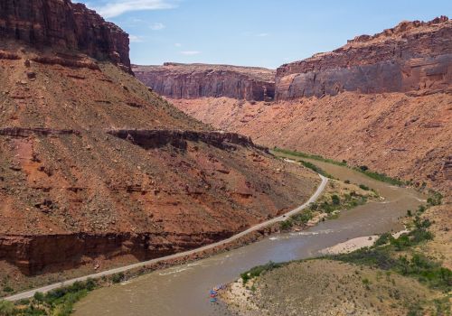 A muddy river winds through a canyon with reddish cliffs and green shrubs, a dirt road tracing along the riverbank in a vast valley.