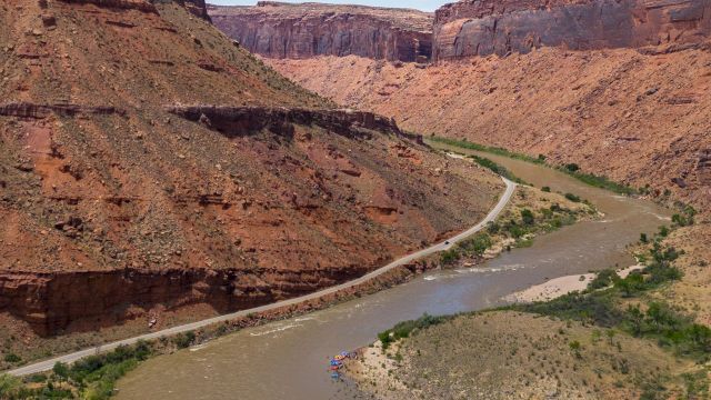 A muddy river winds through a canyon with reddish cliffs and green shrubs, a dirt road tracing along the riverbank in a vast valley.